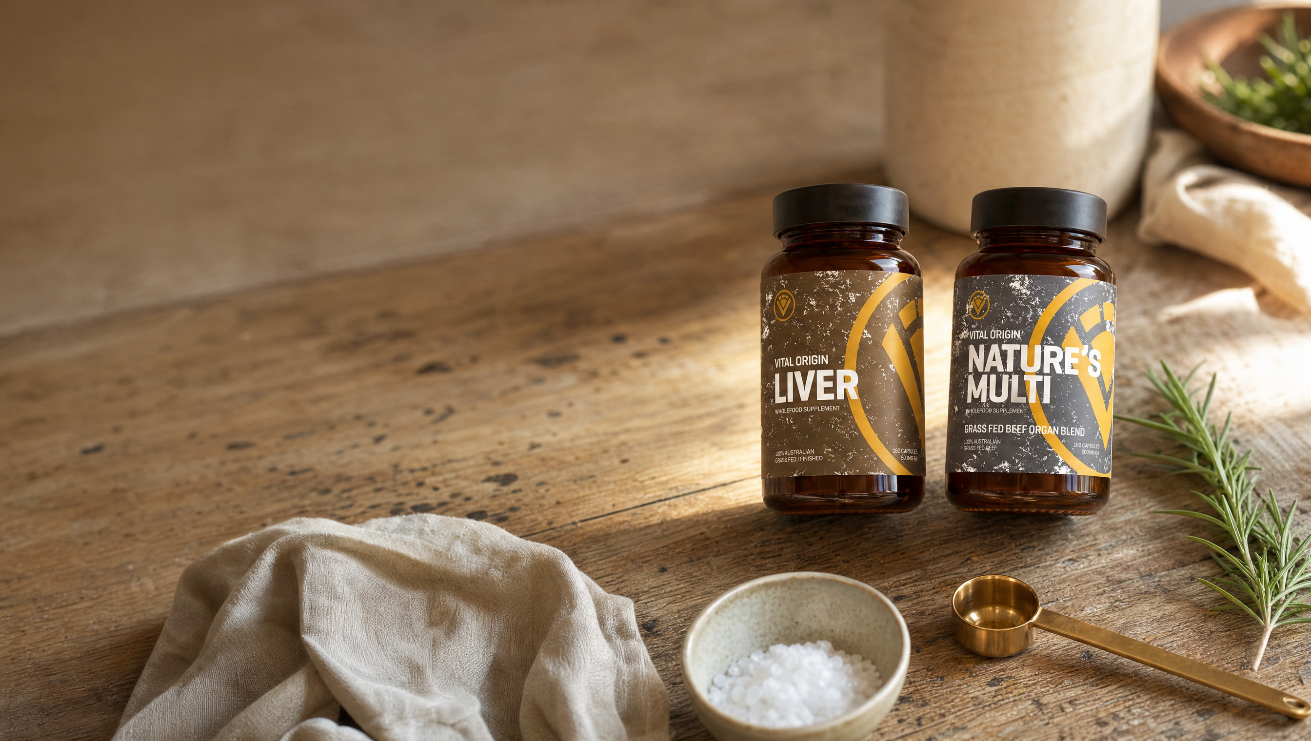 Vital Origin Beef Liver and Nature's Multi wholefood organ supplement tins on a rustic Australian timber kitchen table, illustrating a comparison of processing methods