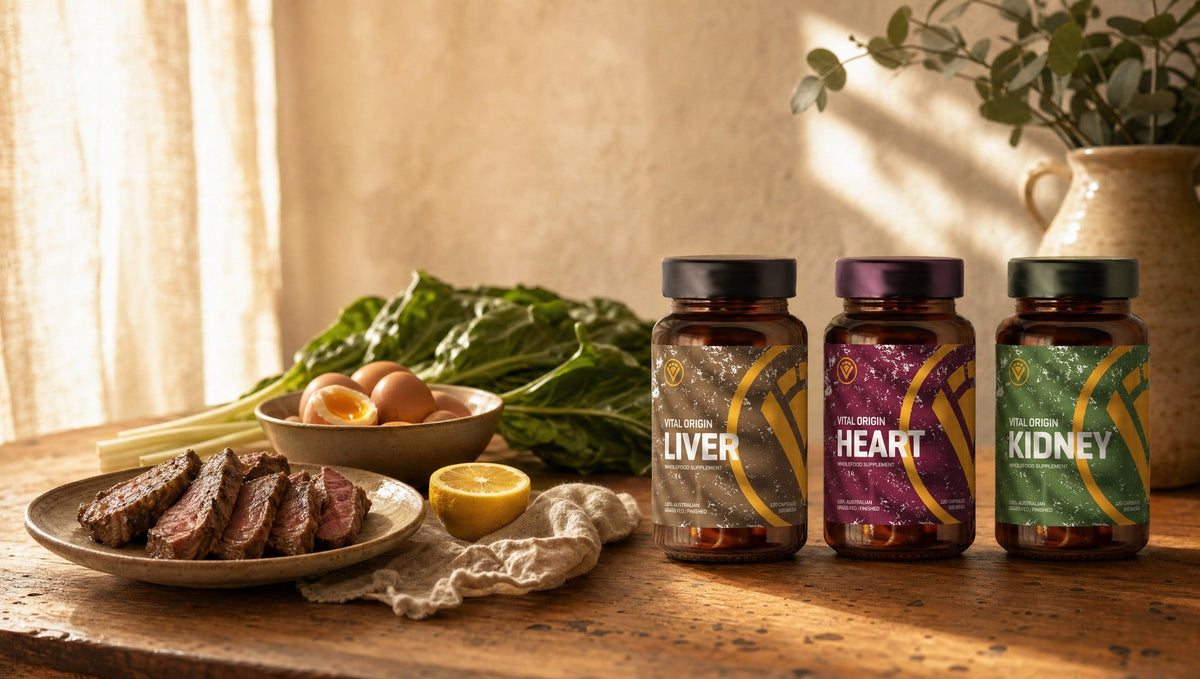 Farmhouse kitchen bench with sliced grass-fed beef, eggs, silverbeet and lemon beside Vital Origin Liver, Heart and Kidney organ capsule bottles in warm morning light
